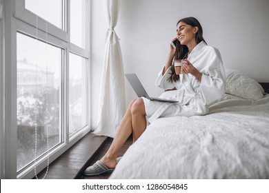 Woman In Bathrobe Sitting In Bed With Computer Drinking Hot Coffee