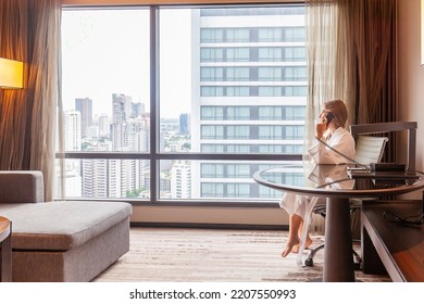 Woman in bathrobe sitting in modern hotel or apartment, talking on phone with city skyscraper view. Holidays in luxury hotel. Businesswoman in hotel room on vacation