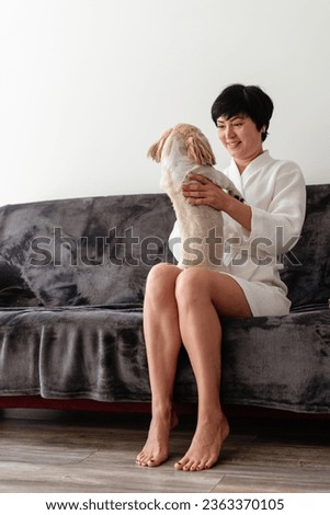 A woman in a bathrobe sitting at sofa and playing, hugging her dog. Brunette woman relaxing at home with her little dog. Love for animals. Good morning concept. Morning routine and domestic lifestyle.