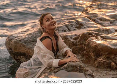 Woman Beach Rocks Sunset - A woman in a white shirt and black swimsuit smiles while sitting on rocks by the ocean at sunset. - Powered by Shutterstock