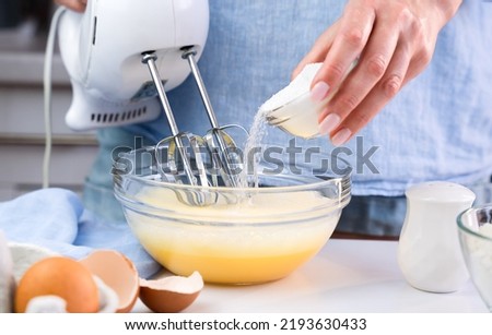 A woman beats eggs with a mixer and adds sugar while standing in the kitchen at home. Cooking and baking desserts at home. Selective focus.
