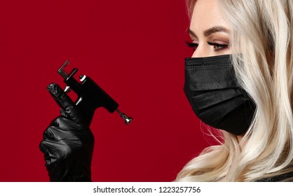 Woman Beautician Cosmetologist Hold New Popular Ear Piercing Gun In Black Medical Gloves And Mask On Red Background