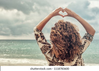 Woman with beautiful curly hair making heart gesture at the sea - Powered by Shutterstock