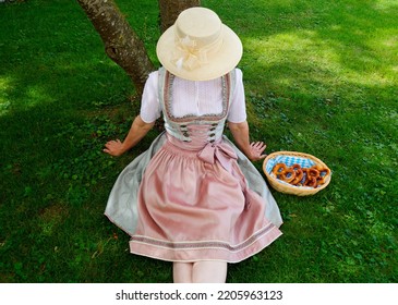 a woman in a beautiful traditional Bavarian or also Austrian dirndl dress sitting on the green meadow with a basket of crunchy salty pretzels or Brezeln (Munich, Bavaria, Germany)                      - Shutterstock ID 2205963123