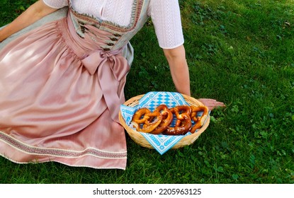 a woman in a beautiful traditional Bavarian or also Austrian dirndl dress sitting on the green meadow with a basket of crunchy salty pretzels or Brezeln (Munich, Bavaria, Germany)                      - Shutterstock ID 2205963125