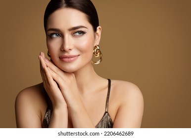 Woman Beauty with Smooth Skin Make up and Golden Jewelry. Beautiful Girl with Perfect Lips and Eye Makeup holding Hands under Chin. Elegant Model Portrait with Gold Earring smiling