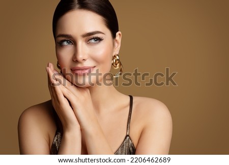 Woman Beauty with Smooth Skin Make up and Golden Jewelry. Beautiful Girl with Perfect Lips and Eye Makeup holding Hands under Chin. Elegant Model Portrait with Gold Earring smiling Foto stock © 