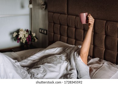 Woman In Bed, Under A Blanket, With Her Hand Raised Up, Holding A Cup Of Coffee. The Morning Ritual Of Waking Up