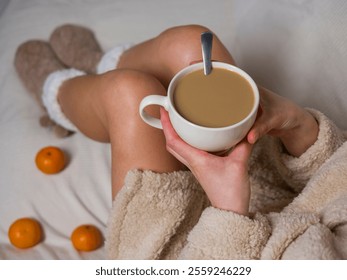 Woman in beige bathrobe and fluffy slippers enjoying a cozy morning at home with a cup of coffee and tangerines in bed - Powered by Shutterstock