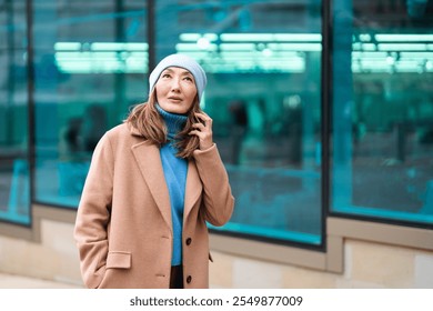 Woman in a beige coat and blue beanie looks up thoughtfully while talking on her phone in an urban setting - Powered by Shutterstock