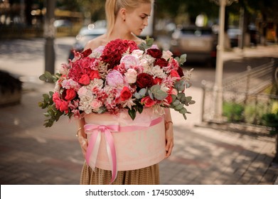 Woman in beige dress holds big bright flower composition with red, pink and white flowers in round box tied with ribbon