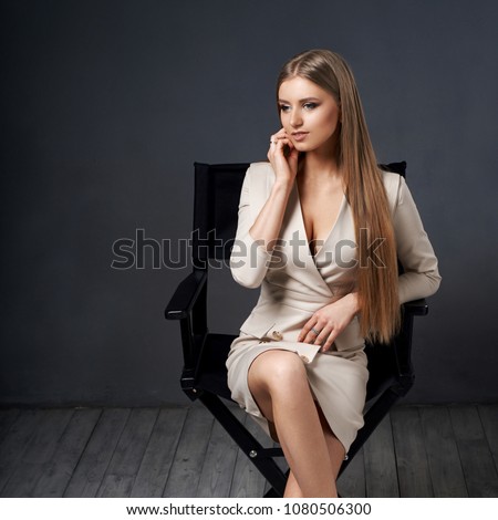 Woman in beige dress jacket sitting at high director's chair in simple interior with wooden floor against gray wall. Girl with long straight blonde hair