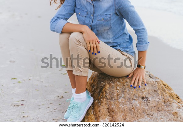 Woman in beige pants and a denim shirt and\
turquoise sneakers sitting on a rock by the sea. Shirt sleeves\
rolled up, watch on her arm, a blue\
manicure.