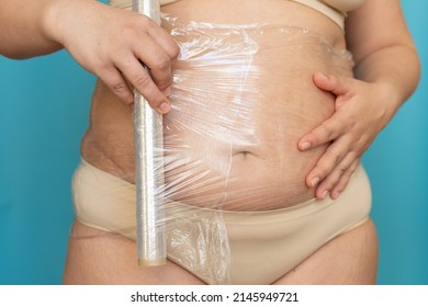 Woman In Beige Underwear Wrap Sagging Belly With Cling Film Closeup. Body Wrapping And Massage For Weight Loss, Free Copy Space, Blue Background. Anti Cellulite Fat Burning Procedure For Slim Figure.