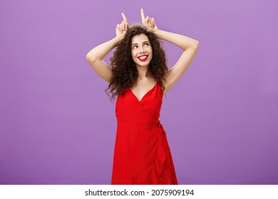 Woman Being Daredevil On Party Standing Sensual And Seductive In Red Elegant Evening Dress Holding Index Fingers On Head Like Devil Horns Looking At Upper Right Corner With Flirty Smile