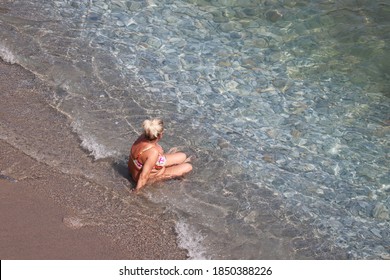 Woman with big breast and fat belly tanning on a beach sitting in sea waves, top view. Vacation, overweight and obesity concept