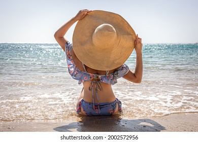 A Woman In A Big Hat Sunbathes On The Beach Near The Sea.