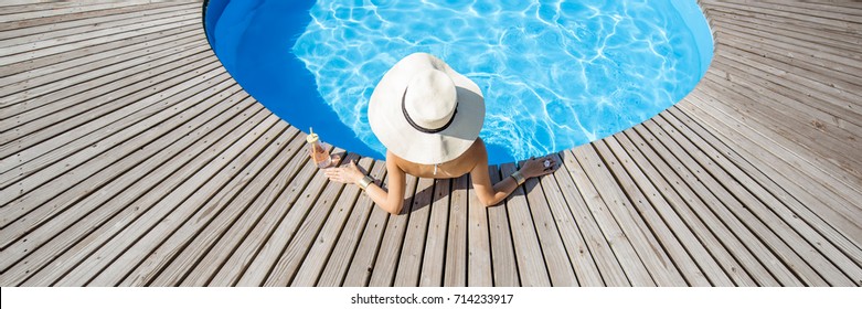 Woman In Big Sunhat With Cocktail Drink Relaxing At The Round Swimming Pool With Blue Water Outdoors. Top View