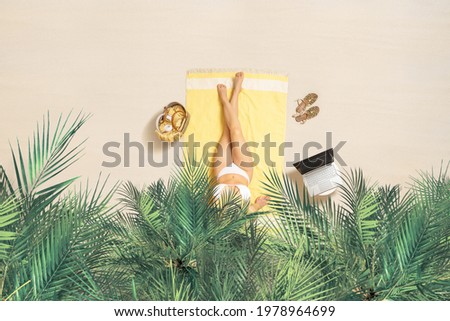 Woman in bikini with laptop, bag and headphones sitting on the beach towel and enjoying summer vacation under palm trees branches. Aerial view