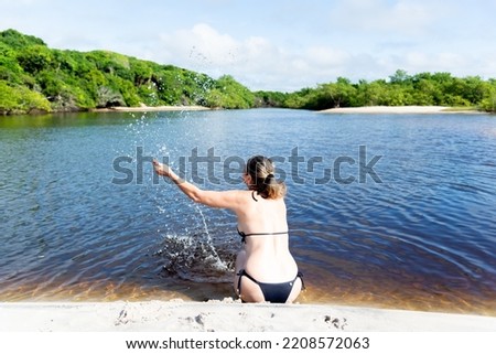 Woman in a bikini sitting on the bank of the river splashes water upwards against the forest and sky in the background. Guaibim beach in the city of Valenca, Bahia, Brazil.