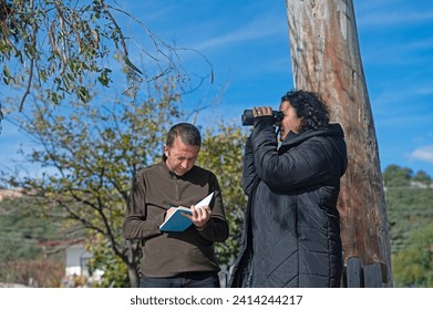 A woman birdwatching with binoculars and a man writing notes in a notebook. - Powered by Shutterstock