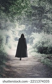 A Woman In A Black Cape Walks Through A Dark, Spooky Forest. Halloween Concept