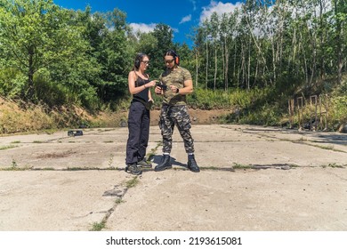 Woman In Black Clothes Showing Man In Camo Clothes How To Operate Handgun. Client With Instructor At Firing Range. Outdoor Horizontal Shot. High Quality Photo