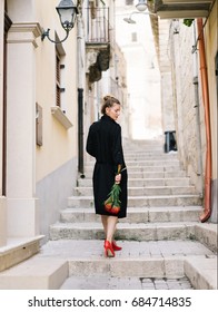 Woman in black coat holding flowers and looking over shoulder on background of old narrow street. 