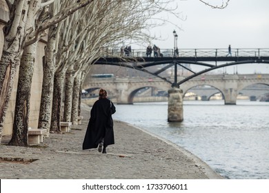 A Woman In A Black Coat Is Walking Along The Seine Embankment.