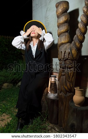 A woman in a black conical hat and black suit with her hands and eyes closed sits next to a wooden Goblin sculpture at night.