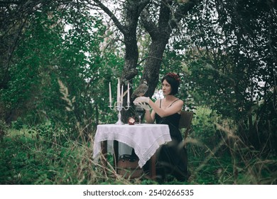 A woman in a black dress with a berry wreath sits at a lace-covered table in a forest, holding a bird while surrounded by lit candles, an apple, and a glass of wine - Powered by Shutterstock