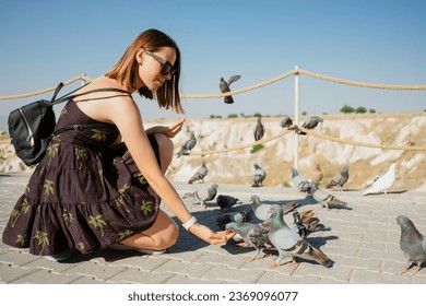 Woman in black dress with green palms on it crouches down to feed pigeons on a stone pavement. The background shows a rope fence, a clear blue sky and a rocky landscape
