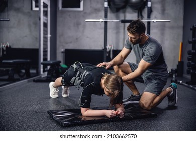 A woman in a black EMS training suit laying in a plank position on her elbows in the middle of a training center. The personal trainer corrects her body position and they prepare to start training
