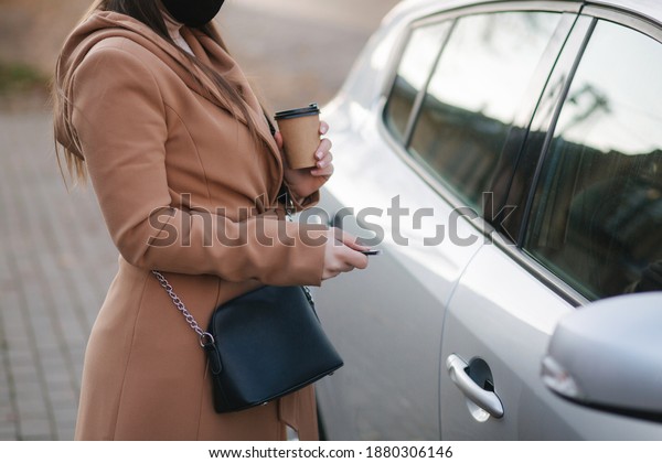 Woman in black face mask hold cuf of coffee by the\
car and look car key. Woman open car. Protectiv mask in quarantine.\
Cov-19