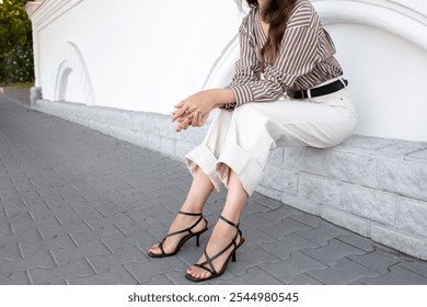 Mujer en sandalias de tacón negro, pantalones de mezclilla blancos y camisa marrón a rayas de seda sentada afuera en la calle de la ciudad. Elegante atuendo casual de moda. Moda callejera. Las piernas de las mujeres, sin rostro.