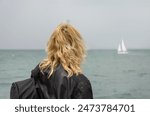 A woman in a black jacket and with a backpack on her shoulder looks at a sailboat at sea