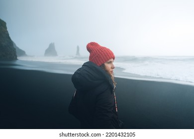 A woman in a black jacket and winter hat is standing on a black beach in Iceland. Hazy rocks and waves in the background. Misty rainy day.