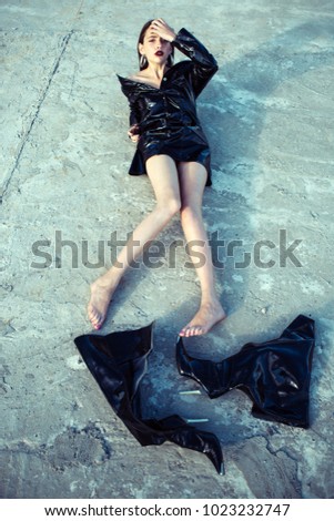 Woman in black latex jacket, barefoot without knee high boots, fashion. Girl with makeup face lie on cement ground, beauty. Fashion, style, accessory. Beauty, look, youth.