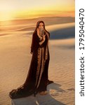 woman in black long dress stands in desert. Luxurious clothes, gold veil accessories hide face. Oriental beauty fashion model. Sand dunes background yellow orange sunset. abaya dress, hijab headscarf