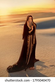 woman in black long dress stands in desert. Luxurious clothes, gold veil accessories hide face. Oriental beauty fashion model. Sand dunes background yellow orange sunset. abaya dress, hijab headscarf