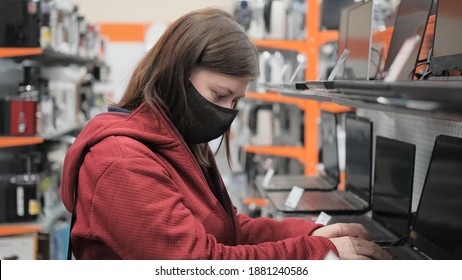 Woman In A Black Mask Against The Virus Buys A Laptop In A Gadget Store. Concept Of Online Communication And Shopping During The Epidemic And Pandemic