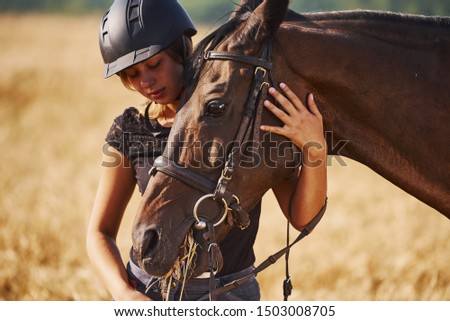 Woman in black protective helmet stands in the field with her horse.