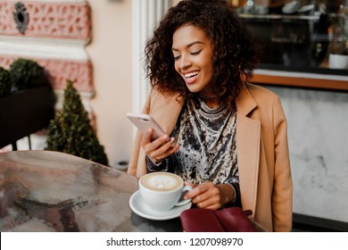 Woman with black skin and candid smile chatting by  phone and enjoying  coffee break in cafe . Wearing elegant velvet dress and beige coat. 