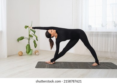 A woman in a black sports jumpsuit practicing yoga performs a variation of the Utthita Trikonasana exercise, an extended triangle pose, training while standing on a mat in the room