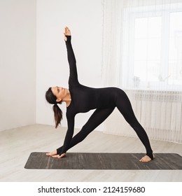 Woman in black sports overalls practicing yoga doing Utthita Trikonasana exercise, extended triangle pose, training while standing on a mat in the room