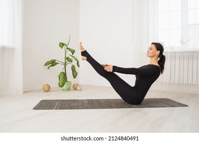 Woman In Black Sports Overalls Practicing Yoga Doing Paripurna Navasana Exercise, Boat Pose, Exercising While Sitting On A Mat In The Room