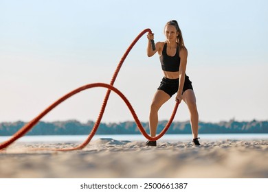 Woman in black sportswear exercising with red battle ropes on a sandy beach, with water and trees in background under clear sky. Concept of sport, active and healthy lifestyle, endurance - Powered by Shutterstock