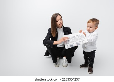 Woman In Black Suit Holding Newspaper Near Little Son While Sitting On Grey