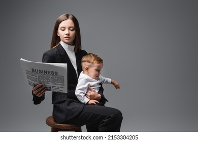 Woman In Black Suit Reading Business Newspaper While Sitting With Son On High Stool Isolated On Grey