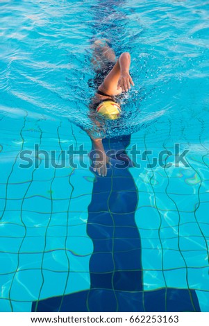 A woman in black swim suits and yellow cap is swimming in freestyle stroke (front crawl) in a swimming pool with clear and blue water.